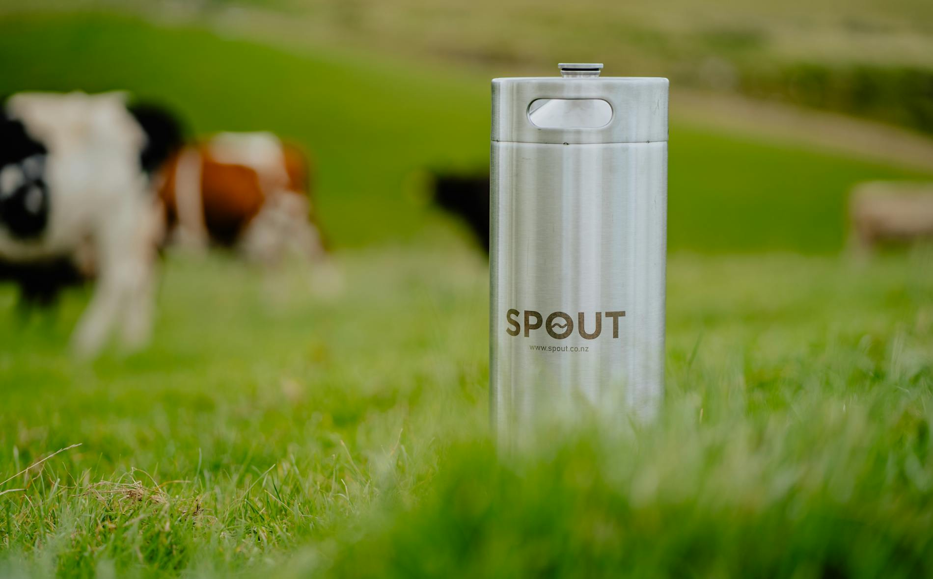 A metallic water container labeled “SPOUT” stands upright on lush green grass, while blurred cows graze peacefully in the background, creating a serene rural ambiance.